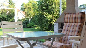 A patio with a glass table and white chairs, a stone chimney, and a view of a house and trees. - Holiday house in the center of Jever - In the house time-out, outside Frisian-tart (Jever)