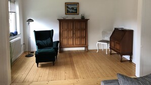 A room with wooden flooring, a green armchair, a white chair, a wooden cabinet, and a floor lamp. - Holiday house in the center of Jever - In the house time-out, outside Frisian-tart (Jever)