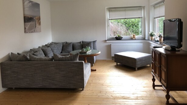 A living room with a grey sofa, a small round table, a television on a wooden cabinet, and a window with blinds. - Holiday house in the center of Jever - In the house time-out, outside Frisian-tart (Jever)