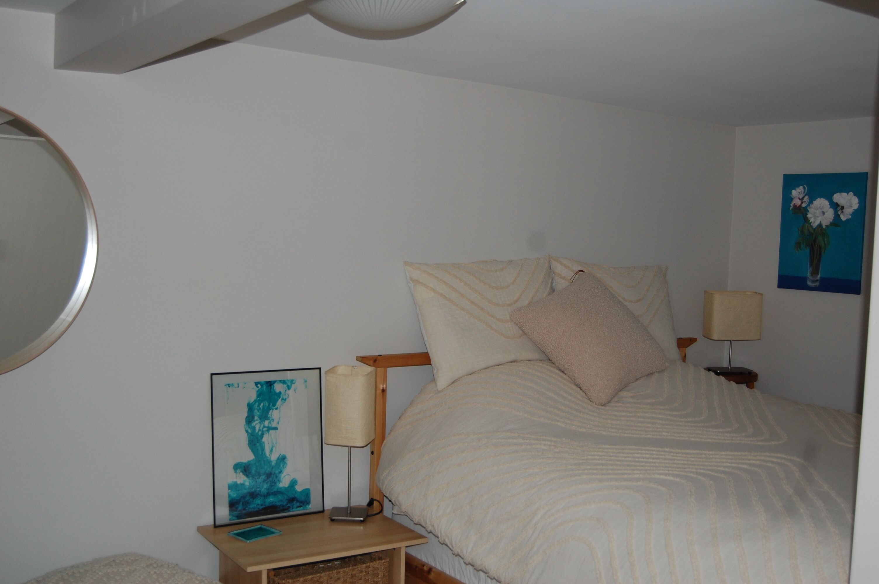 A bedroom with a bed, a bedside table with a lamp, a framed picture, and a wall-mounted mirror.