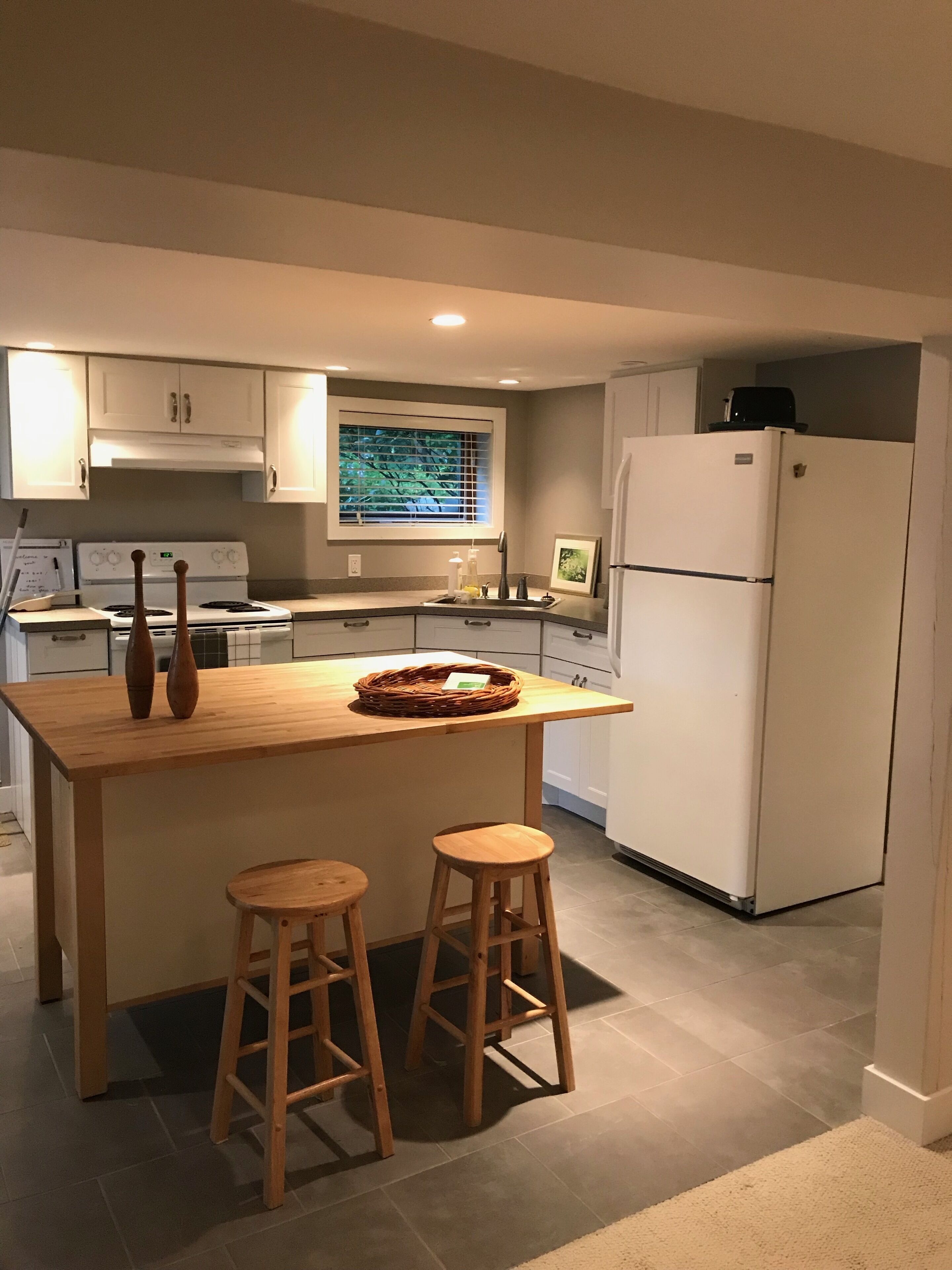 A modern kitchen with a central island, two wooden stools, a refrigerator, and a window with blinds.