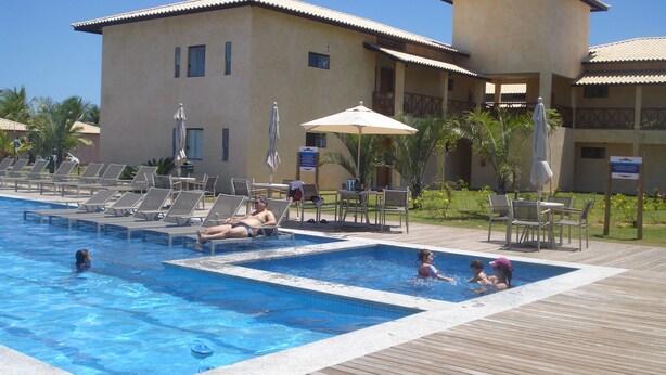 A pool area with lounge chairs and umbrellas, a wooden deck, and a building with a tiled roof. - 3/4 Village in Quinta das Lagoas Reserva - Itacimirim Beach  (Camaçari)