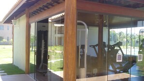 A covered outdoor gym area with exercise equipment, glass walls, and a view of a building and greenery in the background. - 3/4 Village in Quinta das Lagoas Reserva - Itacimirim Beach  (Camaçari)