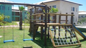 A playground with swings and a climbing structure, surrounded by grass and palm trees, with buildings in the background. - 3/4 Village in Quinta das Lagoas Reserva - Itacimirim Beach  (Camaçari)