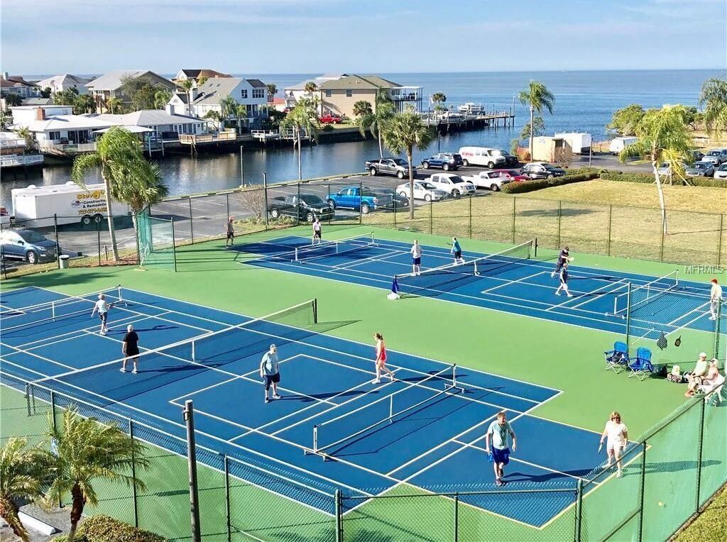 Un court de tennis comprenant plusieurs terrains, entouré d’une clôture, avec vue sur l’océan et des maisons en arrière-plan.