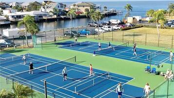 Un court de tennis comprenant plusieurs terrains, entouré d’une clôture, avec vue sur l’océan et des maisons en arrière-plan.