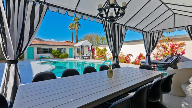 Ein Poolbereich mit Tisch und Stühlen, eine überdachte Sitzgelegenheit und Blick auf ein Haus und Palmen.