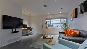 Ein modernes Wohnzimmer mit Flachbildfernseher, Glastisch, Ledersofa mit orangenen Kissen und Essbereich mit Holztisch und Stühlen.