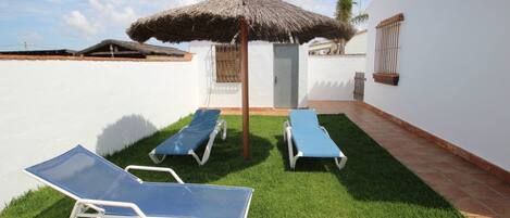 A patio area with two blue sun loungers, a thatched umbrella, and a white wall with a window and a wall-mounted light fixture.