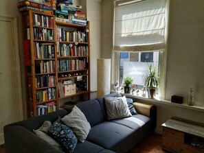 Un salon chaleureux avec un canapé bleu foncé, une bibliothèque en bois remplie de livres et une fenêtre ornée de rideaux légers.