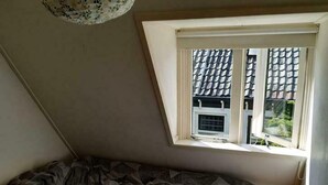 Una habitación pequeña y tenuemente iluminada, con techo inclinado, una cama individual y una ventana que da a un edificio con tejado de tejas.