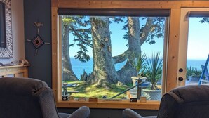 A cozy seating area with two chairs and a small wooden table, a sign reading "BEACH HOUSE", and a view of trees and a beach through a large window. - Ocean Front Home- spacious, beautiful yard, hot tub & ocean view all around!! (Brookings)