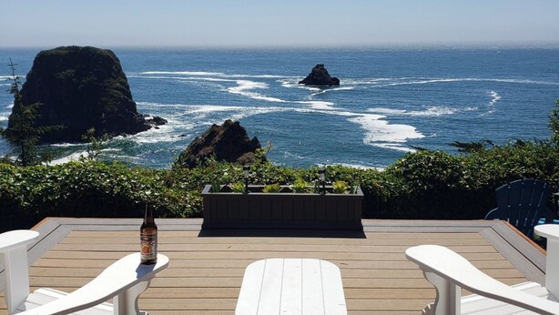 A wooden deck with two white chairs and a small table, overlooking the ocean with waves crashing against rocks. - Ocean Front Home- spacious, beautiful yard, hot tub & ocean view all around!! (Brookings)