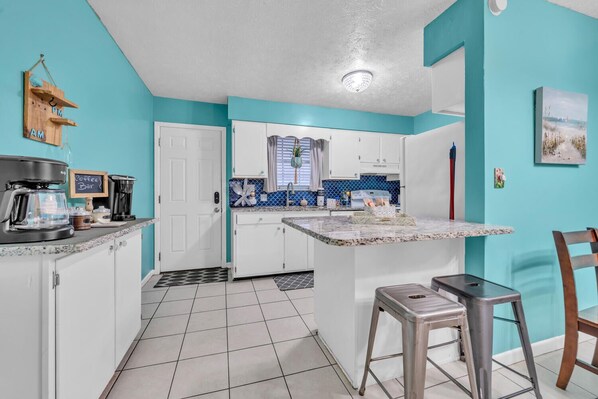 A compact kitchen with white cabinets, a coffee station, and a small dining area with a bar stool.