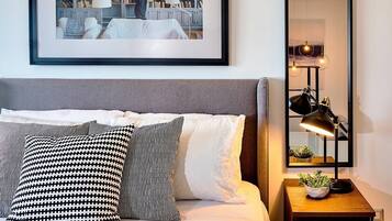 Een keurig opgemaakte slaapkamer met een bed, kussens, een nachtkastje met een lamp, een ingelijst kunstwerk aan de muur en een boekenplank.