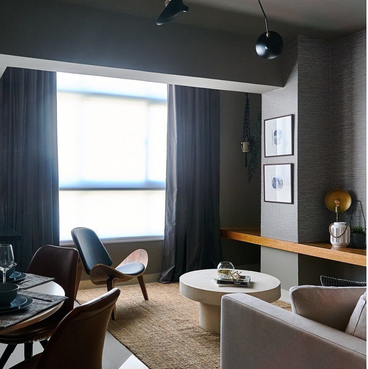 En moderne stue med spiseplads, store vinduer med gardiner, et rundt sofabord og en sofa.