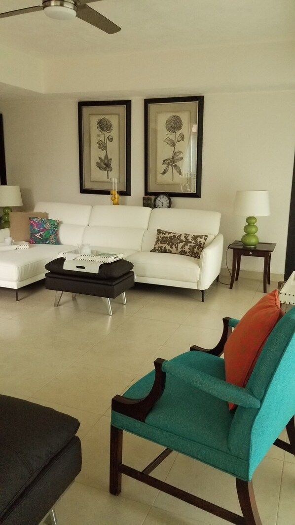 A living room with a white sofa, a teal armchair, a black ottoman, a coffee table, and two framed botanical prints on the wall.