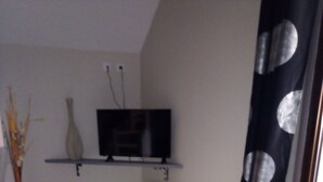 A small, sparsely furnished room with a television on a shelf, a radiator, a white chair, and a window with a curtain.