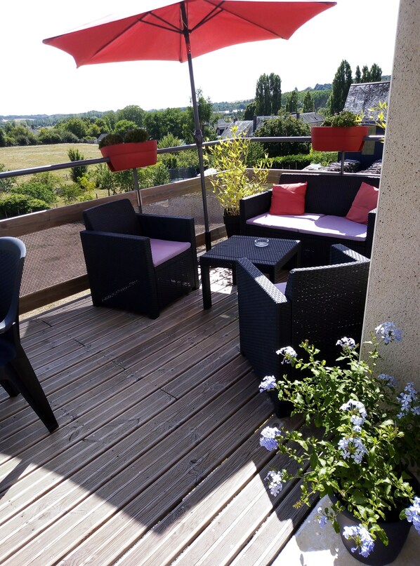 A wooden deck with wicker furniture and a red umbrella, overlooking a green landscape.