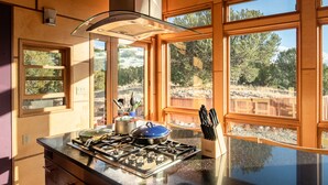 Une cuisine équipée d’une cuisinière à gaz, de casseroles et d’ustensiles, de grandes fenêtres donnant sur la verdure et d’une terrasse en bois à l’extérieur.