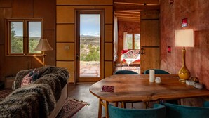 Une pièce chaleureuse avec un plafond en bois, une grande fenêtre, un lit recouvert d’un plaid en fourrure, une table à manger avec des chaises, et une vue sur le paysage désertique.