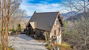 Eine Blockhütte mit Schotterauffahrt, umgeben von Bäumen und Bergen im Hintergrund.