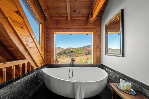 Un baño de cabaña de madera con una bañera independiente, una ventana con vistas a la montaña y un espejo en la pared.