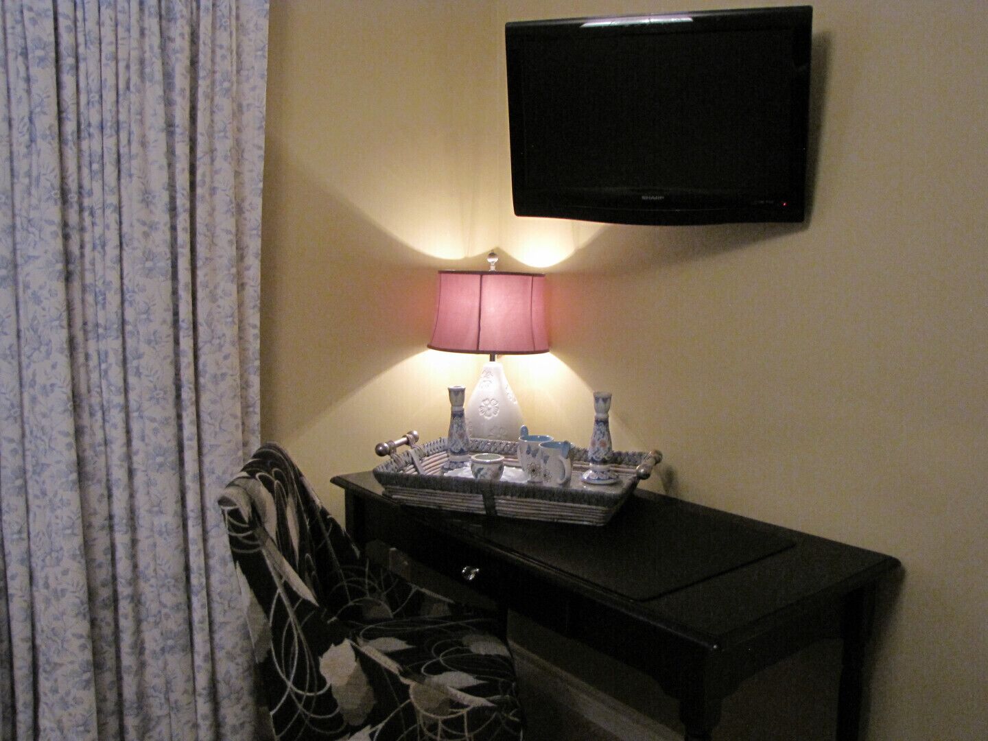 A hotel room with a television mounted on the wall, a bedside table with a lamp and decorative items, and a floral-patterned chair.