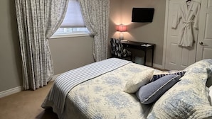 A bedroom with a bed, curtains, a desk, a chair, a robe, and a TV on the wall. - Willow Pond Bed and Breakfast  (Port Perry)