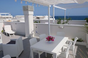 Eine helle Terrasse im Freien mit Tisch und Stühlen, einem weißen Korbsessel und einem Blumentopf mit rosa Blüten.