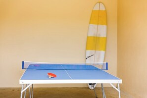 Una mesa de ping pong con una paleta y una tabla de surf apoyada en la pared.