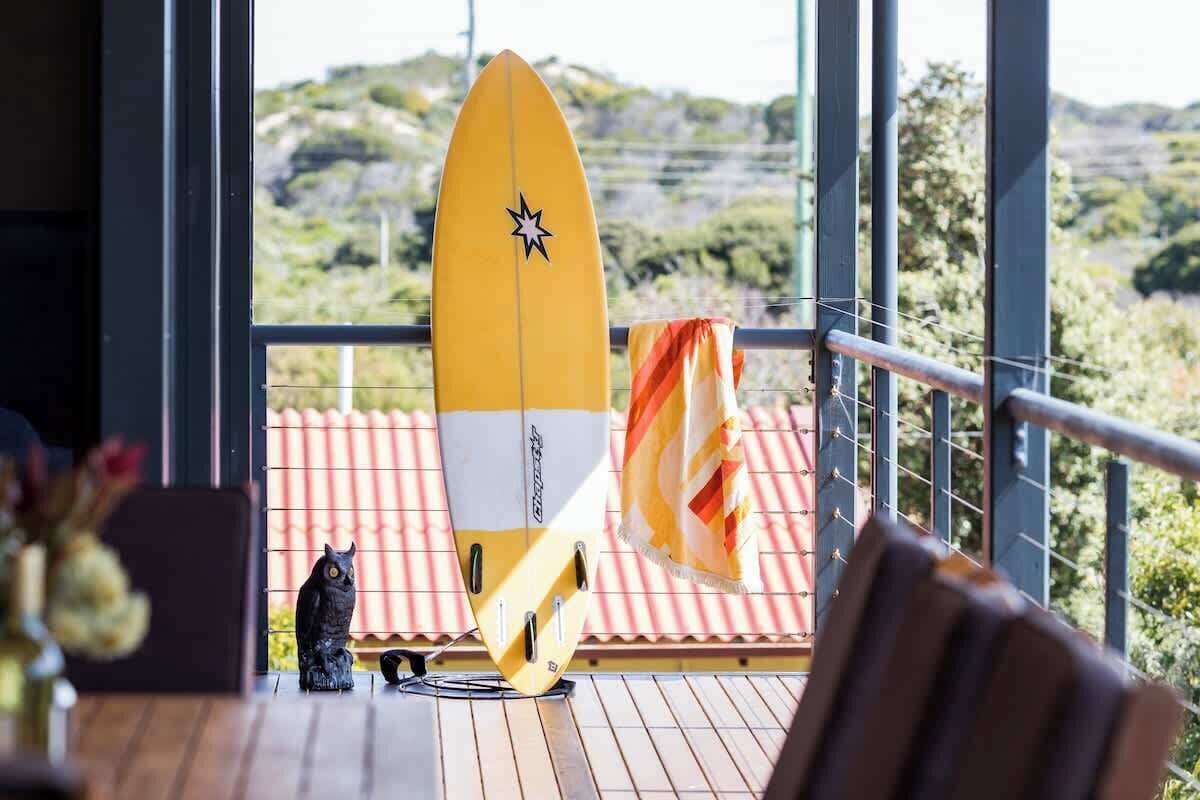 Ein gelbes Surfbrett, eine schwarze Katzenfigur und ein Stuhl auf einem Holzboden mit Blick ins Grüne.