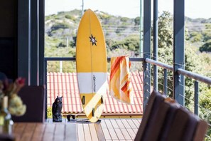 A yellow surfboard, a black cat figurine, and a chair on a wooden deck with a view of greenery.