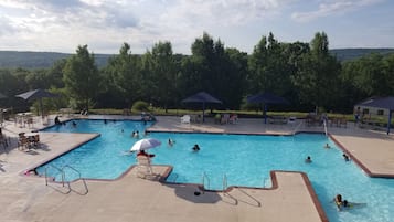 A swimming pool with multiple lanes, surrounded by lounge chairs and umbrellas, with a view of trees and hills in the background.