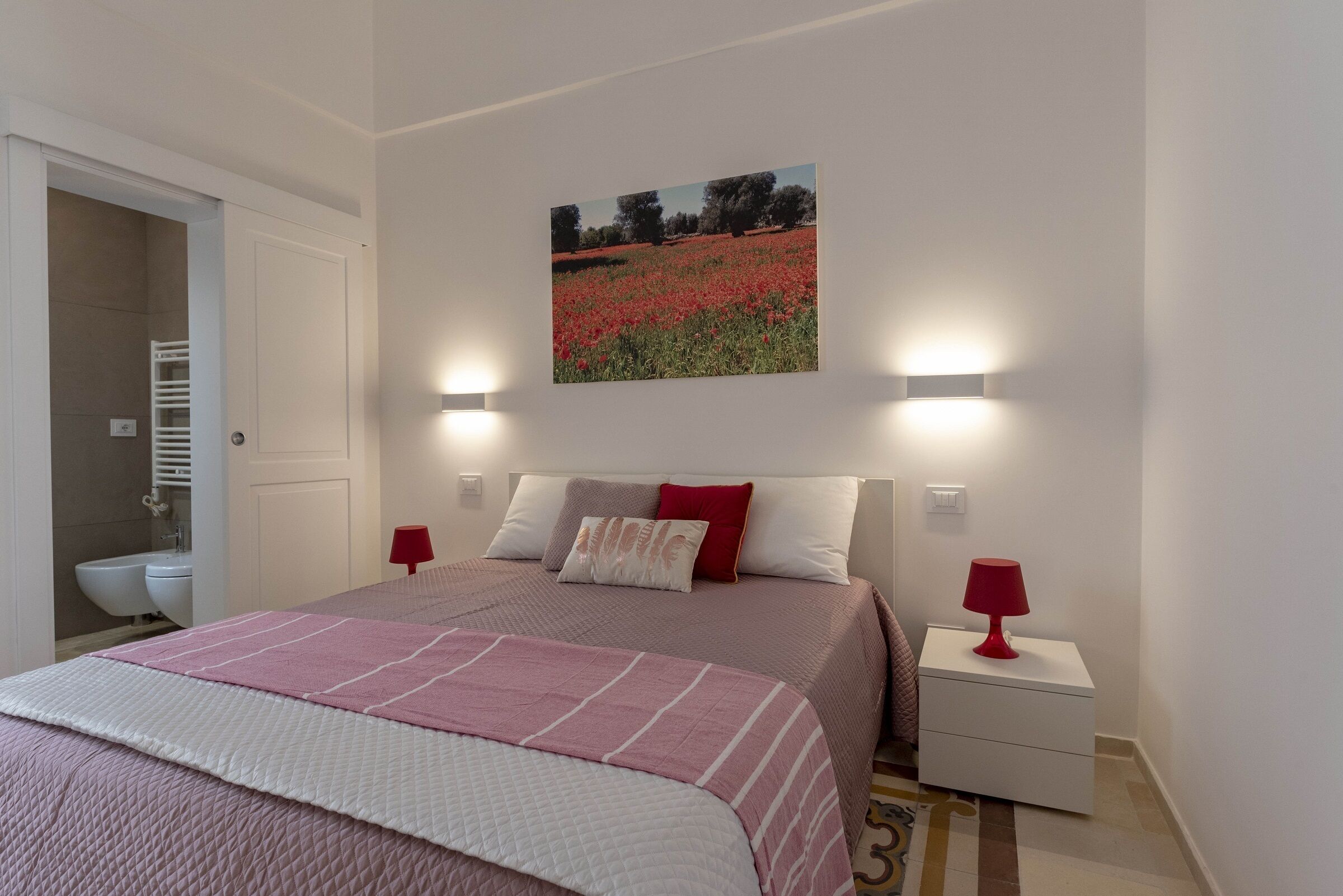 Una camera da letto con un letto, un comodino, una lampada rossa, un quadro di un campo e un bagno con il water e il lavandino.