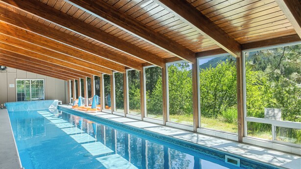 A long indoor swimming pool with wooden beams and large windows offering views of greenery.