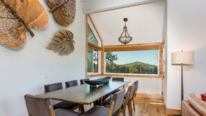 Un comedor con una mesa y sillas de madera, un ventanal con vistas a las colinas y una lámpara colgante.