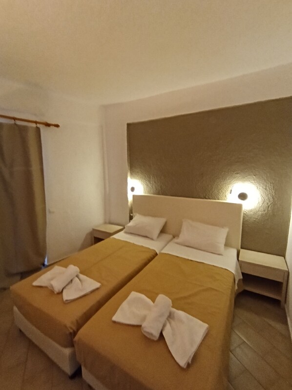 A hotel room with two beds, each with a brown bedspread and white towels folded in a bow. There are two bedside tables with lamps, and the room has a simple, clean design with white walls and a wooden headboard. - Relax vacation for all (Ag. Nikolaos)