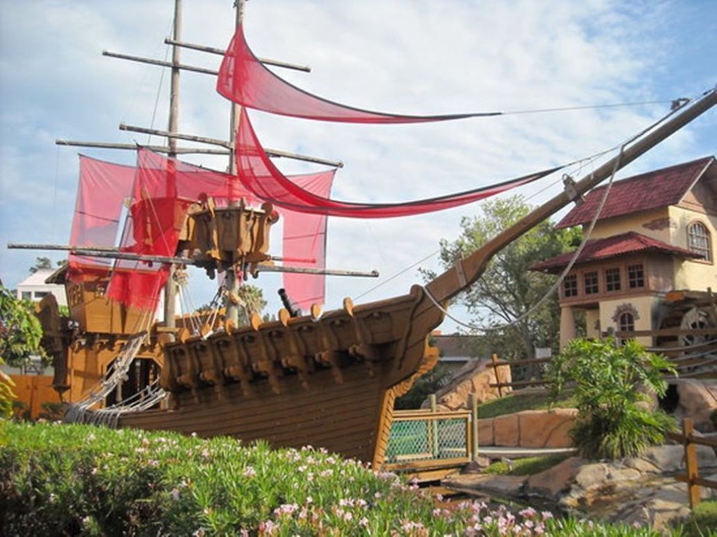 A model ship with a red sail, surrounded by greenery and a building with a red roof.