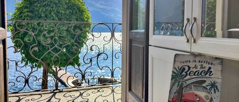 A balcony with a view of the sea and mountains, a metal railing, a potted tree, and a decorative sign.
