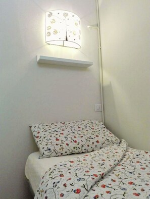 A small room with a bed, a wall-mounted lamp, and a white shelf.