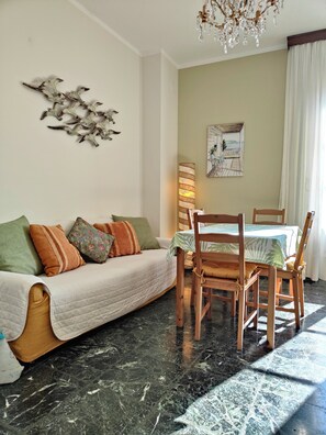 A living room with a sofa, a dining table with chairs, a chandelier, and a wall-mounted artwork.