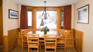Een eetruimte met een houten tafel en stoelen, een kroonluchter en uitzicht door grote ramen.