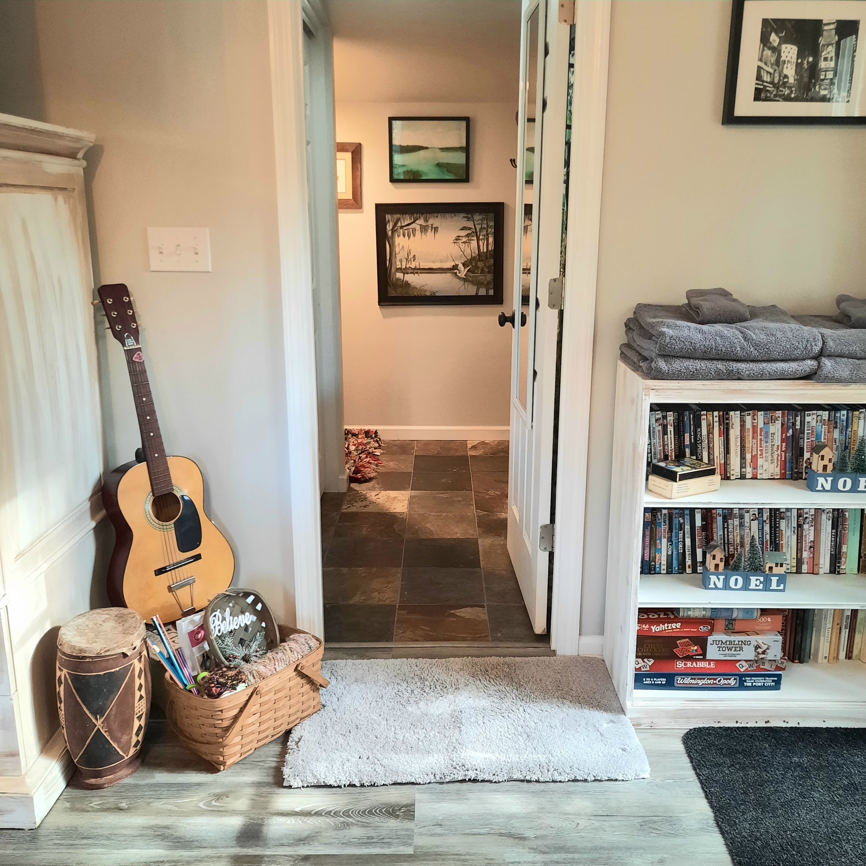 A hallway with a guitar, a drum, and a basket of items. There is a bookshelf with books and a 'NOEL' decoration.