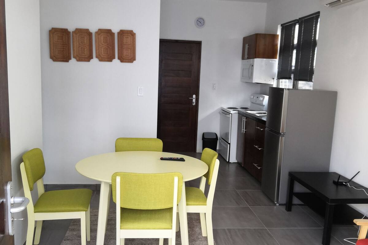 A compact living space with a dining area featuring a round table and four chairs, a kitchen with modern appliances, and a small black table with a laptop.