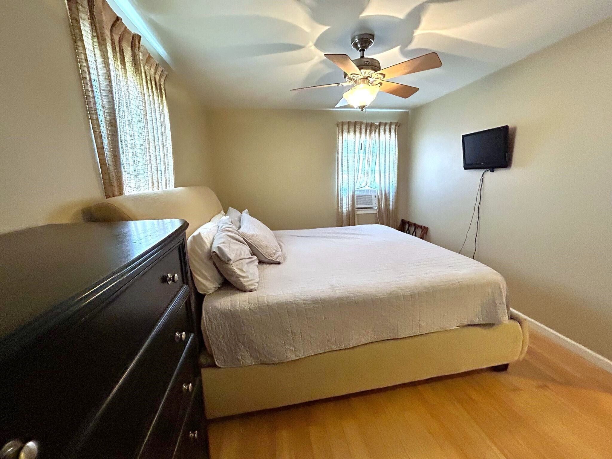 A bedroom with a bed, a dresser, a ceiling fan, a TV, and a window with curtains.