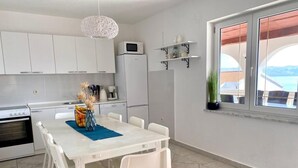 A modern kitchen with white cabinets, a dining table with chairs, a hanging light fixture, and a view of the sea through a window. - Villa Mihaela - Beachfront apartment "Mario" with sea view and pool (Cesarica)
