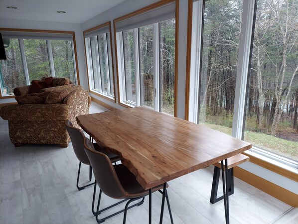 A modern dining area with a wooden table and chairs, large windows with a forest view, and a patterned sofa.