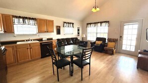 A spacious living area with a dining table and chairs, a kitchen with wooden cabinets, and a large window providing natural light. - La Bon Vie - Coastal Realty Group (Grand Isle)