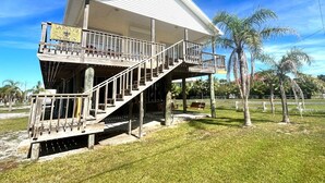 A two-story building with a wooden deck and a balcony, surrounded by palm trees and a clear blue sky. - La Bon Vie - Coastal Realty Group (Grand Isle)